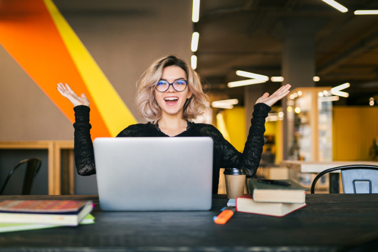 funny happy excited woman sitting at table in black shirt working on laptop in co-working office, wearing glasses