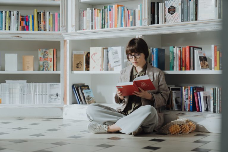 Mergina sėdi ant žemės ir skaito knygą knygų lentynų fone