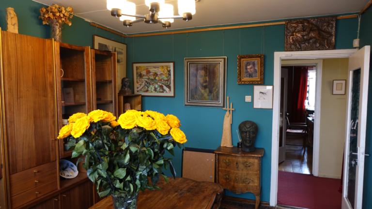 Knygų lentyna, ant sienos kabo paveikslai, ant stalo stovi rožių puokštė, durys į kitą kambarį