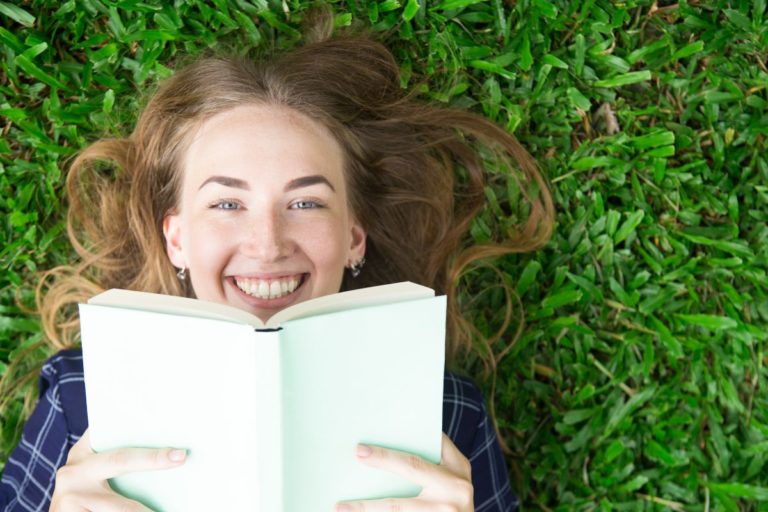 Mergina guli ant žolės, šypsosi ir laiko knygą