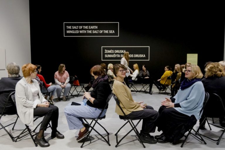 Grupė moterų sėdi ant kėdžių po dvi viena prieš kitą muziejaus aplinkoje