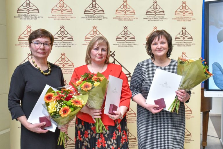 Trys moterys šypsosi su gėlių puokštėmis ir apdovanojimais