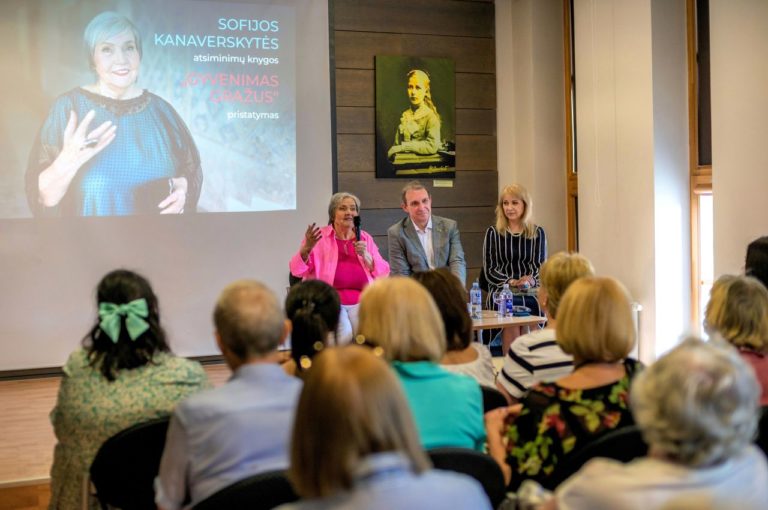 Trys žmonės sėdi ant pakylos prieš gausią žmonių auditoriją