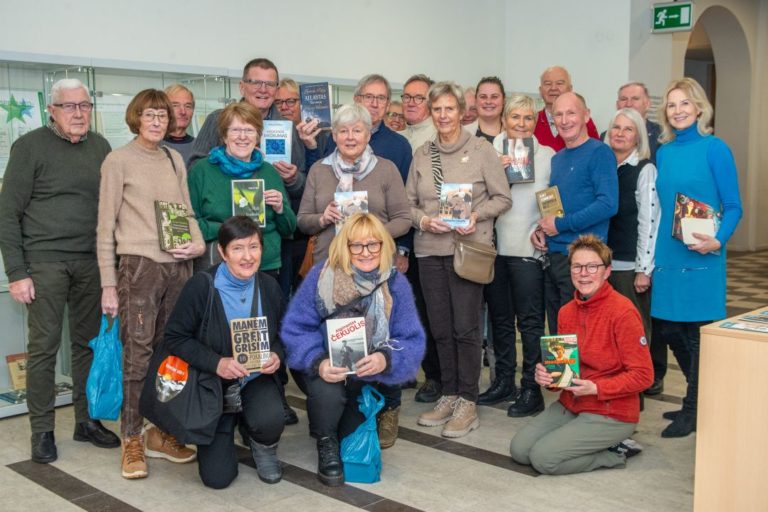 Būrys besišypsančių žmonių su knygomis rankose bibliotekos aplinkoje