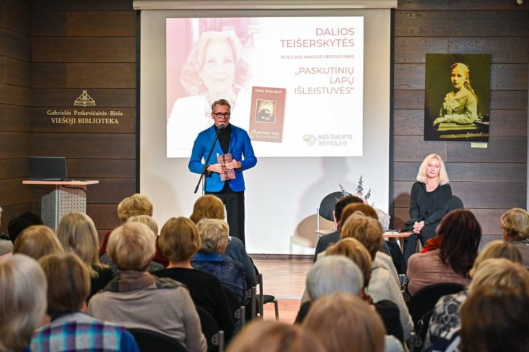Vyras su knyga rankoje stovi priešais renginio dalyvių auditoriją, šalia veidu į auditoriją sėdi moteris.