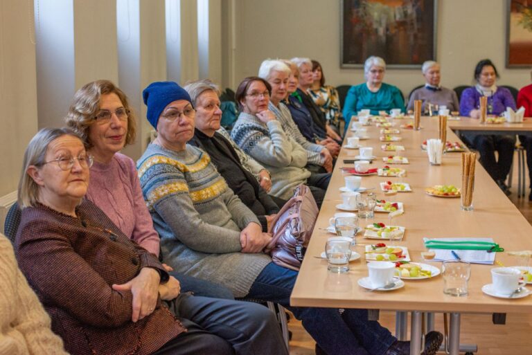 Didelė grupė žmonių sėdi prie ilgo stalo su indais ir maisto produktais