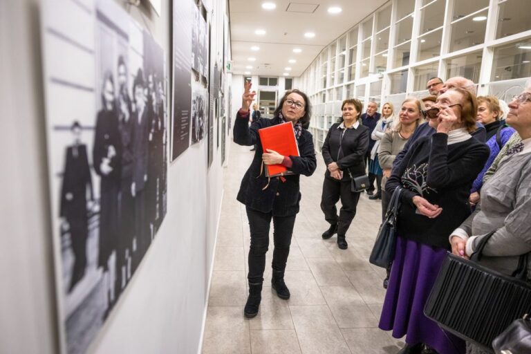 Moteris pasakoja prie parodos nuotraukų pakėlusi ranką grupei klausytojų bibliotekos erdvės fone
