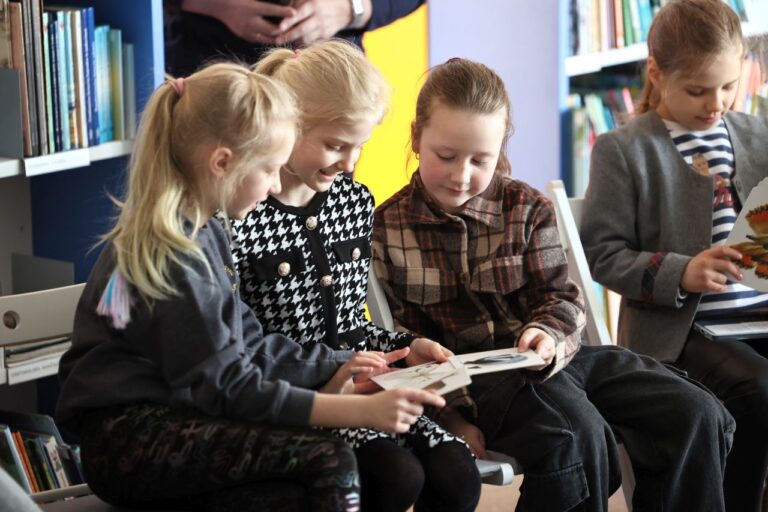Jaunesnio amžiaus trys mergaitės sėdi bibliotekos aplinkoje, žiūri į paveiksliuką ir šypsosi