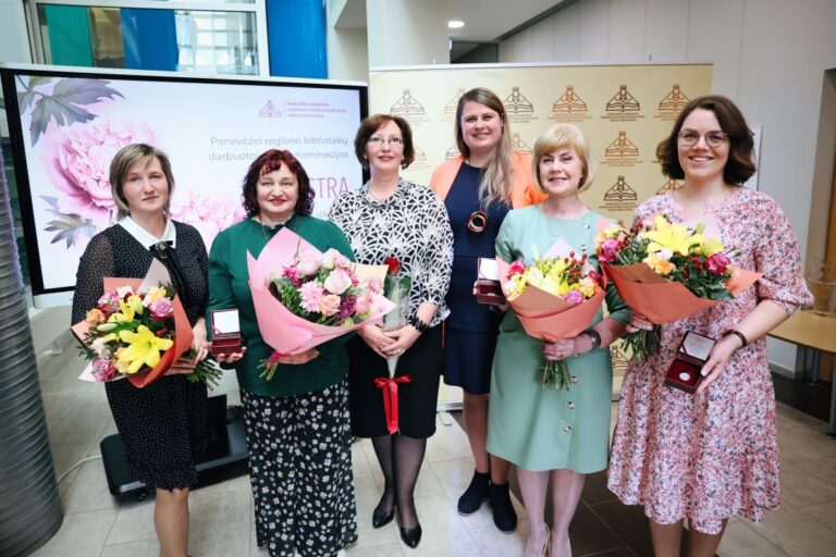 Stovi ir šypsosi šešios moterys, trys iš jų su gėlėmis ir apdovanojimo ženklais rankose bibliotekos erdvėje