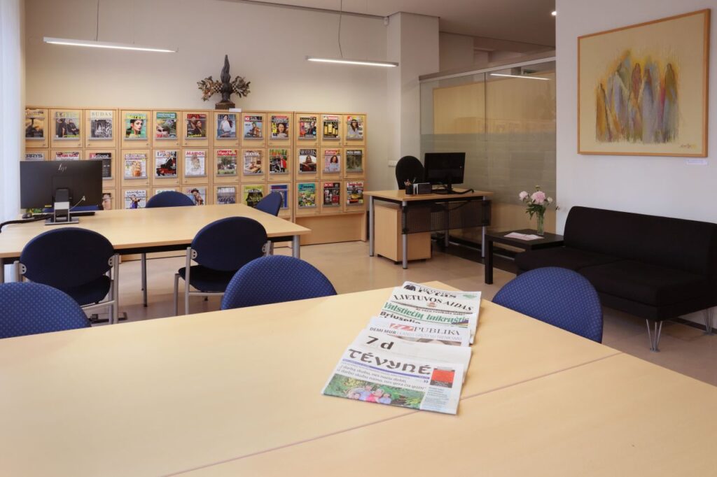 Bibliotekos skaityklos erdvėje žurnalų lentynos, stalai su kėdėmis, ant stalo išdėlioti laikraščiai