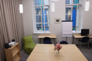 Kvadratinis stalas centre, šonuose du mažesni stalai, rašymo lenta, projektorius patalpos langų fone