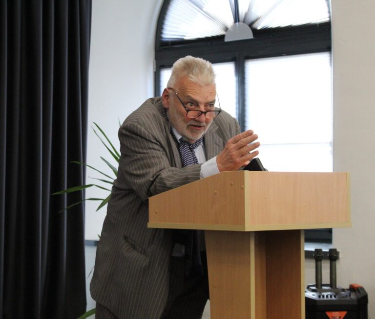 Vyras žilais plaukais kalba priešais auditoriją tribūnoje salės erdvėje