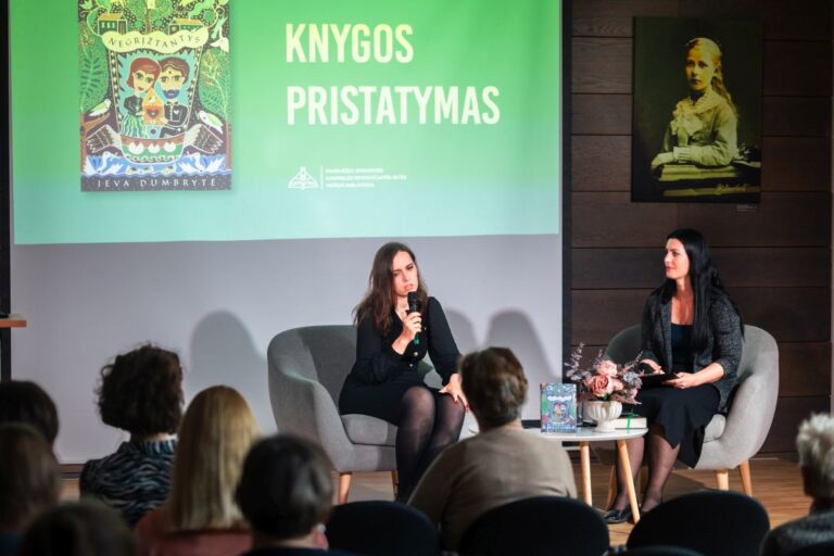 Dvi moterys kalbasi sėdėdamos foteliuose ant pakylos priešais renginio dalyvių auditoriją salės fone
