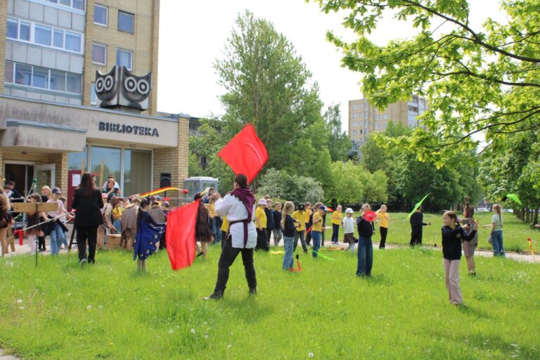 Grupė vaikų užsiima įvairiomis aktyviomis veiklomis lauke bibliotekos fone