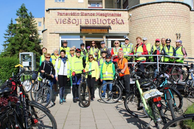 Didelė grupė žmonių su ryškiomis liemenėmis stovi prie įėjimo į biblioteką dviračių apsupty