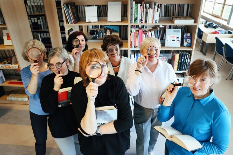 Būrelis moterų pridėję didinamuosius stiklus prie akių bibliotekos knygų lentynų fone