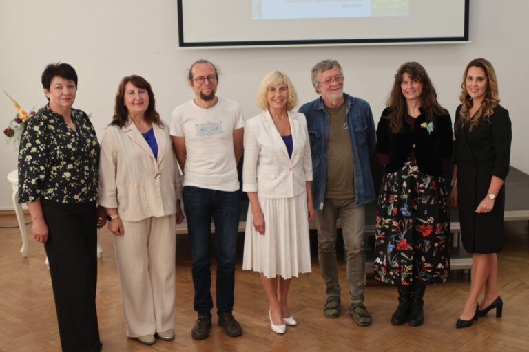 Grupė žmonių vyrų ir moterų stovi visu ūgiu priešais renginio auditoriją