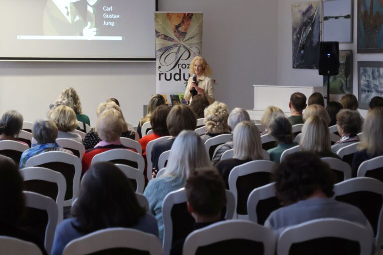 Tradicinis „Prozos ruduo“ su rašytoja Eva Tombak Moteris stovi priešais renginio dalyvių auditoriją salės fone