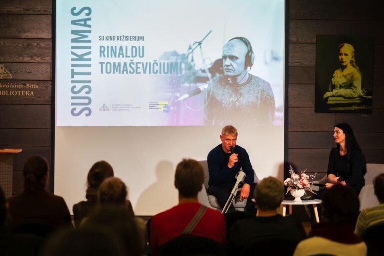 Rinaldo Tomaševičiaus pasakos kaip gyvenimas Renginio svečias režisierisu su vedėja sėdi ant pakylos priešais renginio auditoriją ir kalbasi salės fone