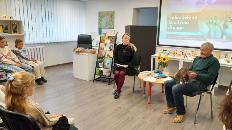 Priešais vaikų auditoriją sėdi moteris ir vyras, kuriam ant kelių tupi katė salės fone