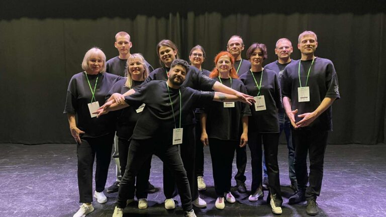 Grupinė nuotrauka scenoje: ant juodo fono stovi vienuolika žmonių, apsirengusių juodais marškinėliais ir avinčių sportinius batelius. Visi su žaliais virveliniais identifikaciniais ženkliukais. Priekyje viduryje vienas žmogus plačiai išskėtęs rankas ir šypsosi, o kiti stovi už jo dviejose eilėse, taip pat šypsosi.