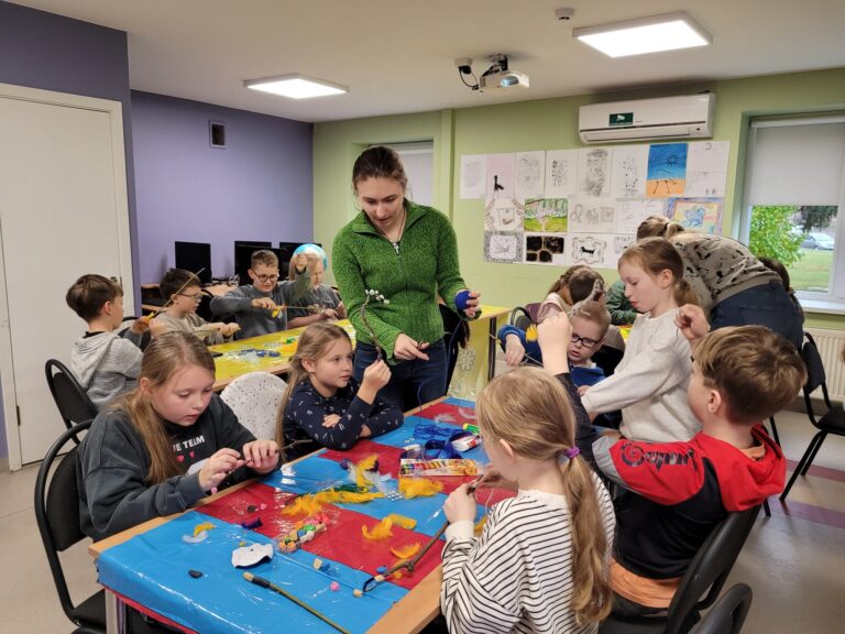 Nuotraukoje matyti grupė vaikų, kurie užsiima kūrybine veikla klasėje. Jie sėdi aplink stalus, padengtus spalvotais plastikiniais užvalkalais, ir dirba su įvairiais amatų reikmenimis, tokiais kaip siūlai, plunksnos ir spalvota molis. Jauna suaugusioji, greičiausiai mokytoja arba dirbtuvėlių vadovė, padeda vaikams ir teikia instrukcijas.