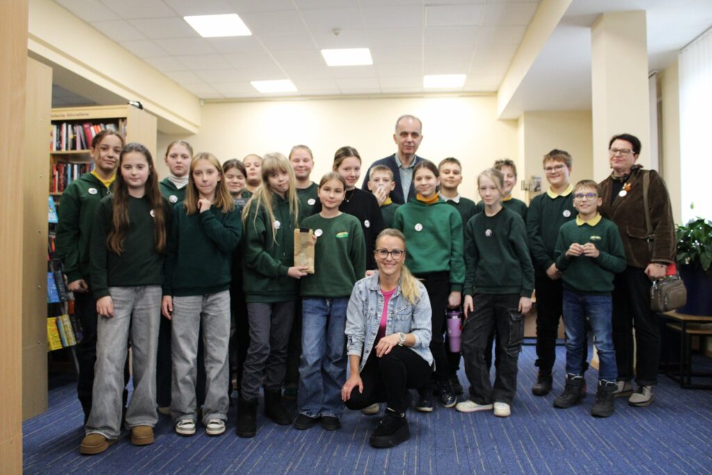 Grupė vaikų ir keli suaugusieji pozuoja bendrai nuotraukai bibliotekoje; vaikai vilki žalias uniformas, o priekyje pritūpusi Aušrinė Tilindė su džinsine striuke šypsosi.