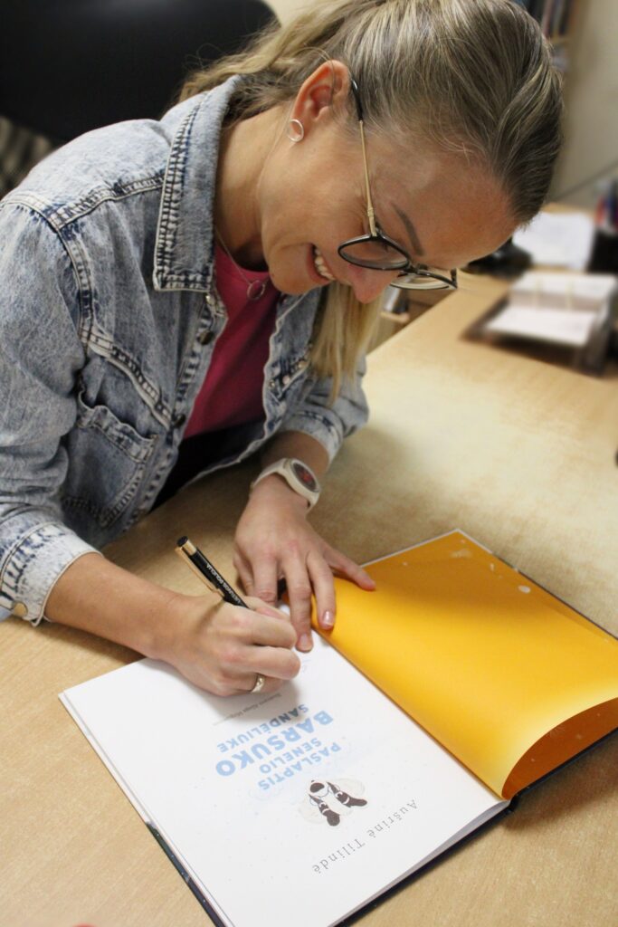 Aušrinė Tilindė sėdi prie stalo ir su šypsena pasirašo Aušrinės Tilindės knygą „Paslaptys senelio Barsuko sandėluike", kurios atverstas puslapis iliustruotas barsuku.