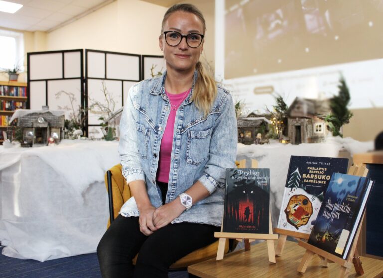 Nuotraukoje matoma autorė, sėdinti prie stalo su savo knygomis. Ji dėvi džinsinę striukę, rožinį marškinėlį ir turi šviesius plaukus, susirištus į uodegą. Už jos yra dekoracija su mažais sniegu padengtais namukais, o priekyje stovi trys jos knygos, kurios yra išdėstytos ant mažų stelažų. Knygos pavadinimai matomi aiškiai, o autorė žiūri į kamerą su šypsena. Fone galima pamatyti bibliotekos ar panašios erdvės detales, kurios sukuria šiltą ir jaukią atmosferą.