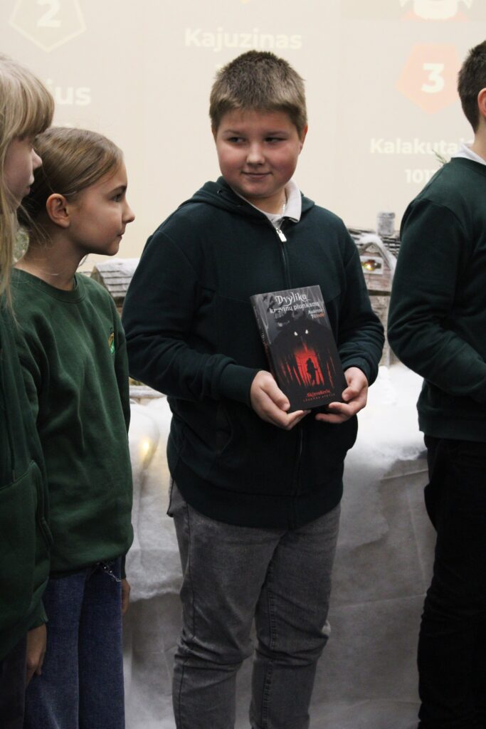 Grupė vaikų stovi kartu bibliotekoje. Centre esantis berniukas laiko Aušrinės Tilindės knygą „Dvylika kruvinų plunksnų“ tamsiu viršeliu. Visi vaikai vilki tamsiai žalias arba tamsias sportinio stiliaus bliuzonas, o fone matyti dekoracijos ir projekcijos su tekstu.