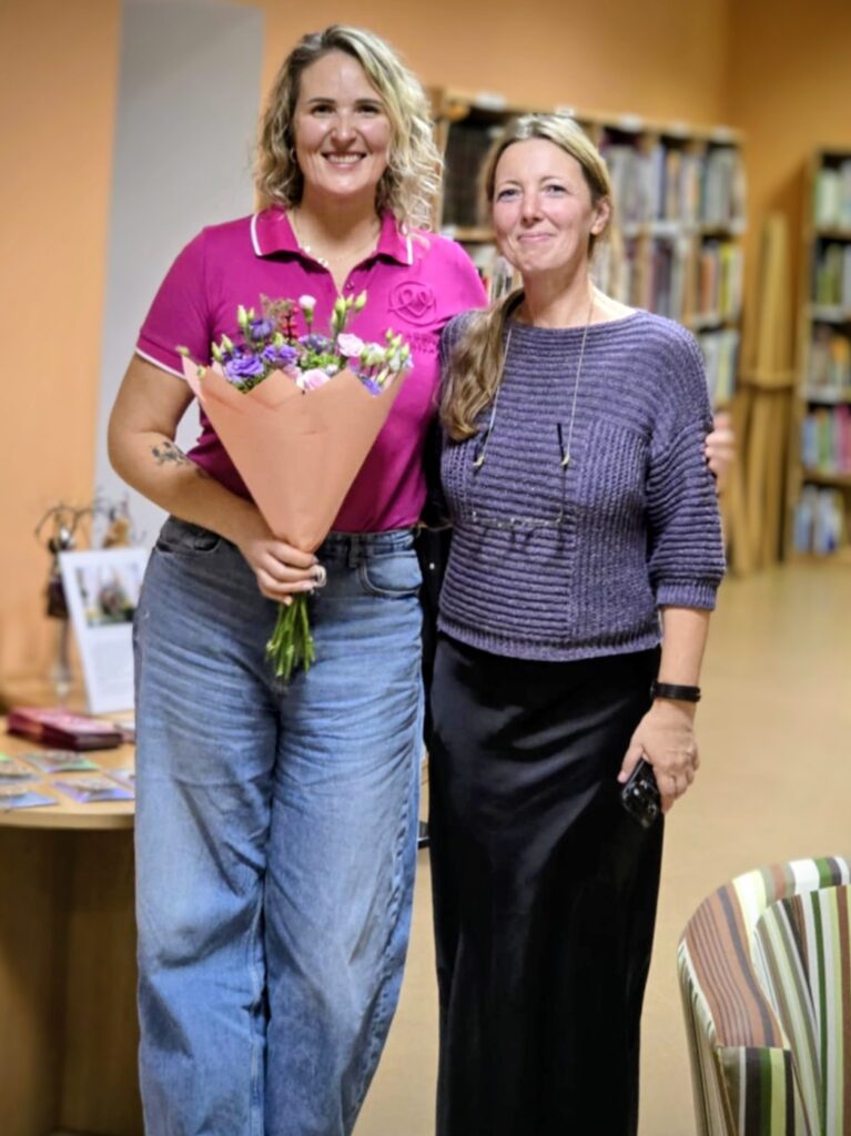 Dvi moterys stovi bibliotekoje: viena vilki rožinius marškinėlius ir laiko gėlių puokštę, kita – violetinį megztinį ir juodą sijoną; jos šypsosi kamerai.