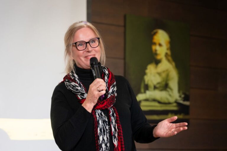 Moteris šypsodamasi kalba į mikrofoną bibliotekos salės fone