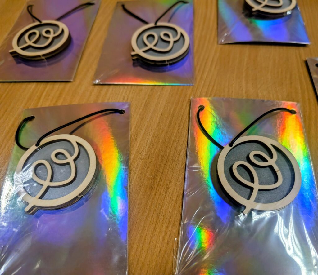 Ant medinio stalo išdėlioti keli pakabukai su pintu linijiniu simboliu, pritvirtinti prie blizgių, holografinių pagrindų.