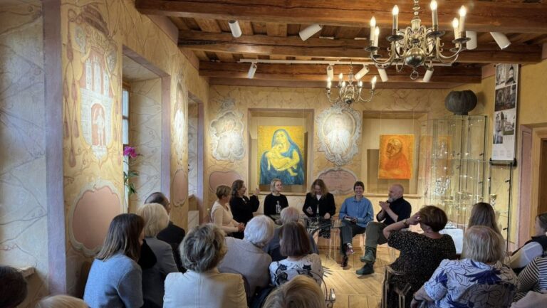 Grupė renginio dalyvių sėdi priešais klausytojų auditoriją vienuolyno salės fone