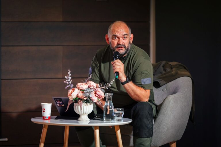 Vyras sėdi ir kalba į mikrofoną salės fone