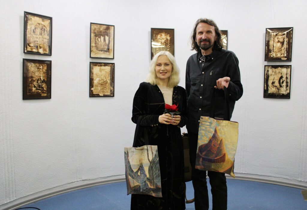 Nuotraukoje matomi du žmonės, kurie pozuoja priešais kelis įrėmintus meno kūrinius, kabojančius ant sienos. Moteris laikosi mažą gėlių kompoziciją rankose, abu laiko Čiurlionio paveikslų motyvais išpieštus pirkinių maišus. Abu dėvi juodus drabužius. Už jų esančioje galerijos erdvėje kabantys kūriniai atrodo abstraktūs arba siurrealistiniai, vyrauja sepijos atspalviai su įvairiais formų ir linijų deriniais, galbūt vaizduojančiais laivus ar architektūrinius elementus.