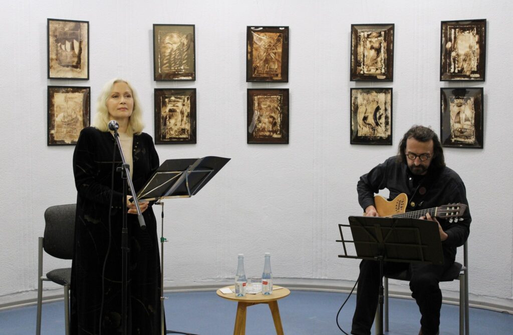Nuotraukoje matyti dvi asmenybės meno parodoje, kuri vyksta prieš meno kūrinius. Moters figūra yra kairėje pusėje, ji stovi prie mikrofono ir laikosi muzikos partiturą, vilkėdama juodą aksominį rūbą. Dešinėje pusėje, ant kėdės, sėdi vyras, grojantis gitara. Ant stalo tarp jų yra du vandens buteliai ir kiti smulkūs daiktai. Sienoje kabantys meno kūriniai turi šiltus atspalvius, panašius į sepiją.