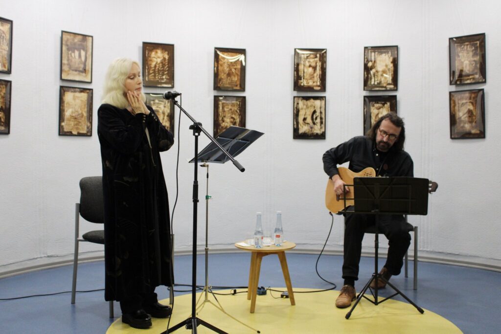 Nuotraukoje matyti moteris, stovinti prie mikrofono, laikanti rankas ant veido, o šalia jos sėdi vyras, grojantis gitara. Moteris vilki juodą aksominį rūbą, o vyras - juodą marškinį ir rudus batus. Ant stalo tarp jų matosi du vandens buteliai ir kiti smulkūs daiktai. Sienose pakabinti meno kūriniai, kuriuose dominuoja šiltos, sepijos spalvos atspalviai. Tai meno ir muzikos renginio akimirka galerijoje.