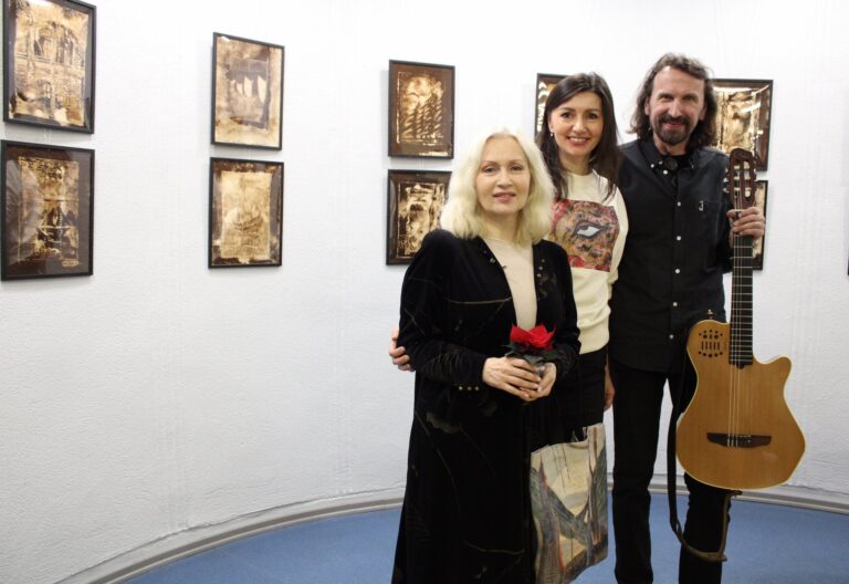 Nuotraukoje matyti trys žmonės, stovintys kartu prieš meno parodą. Kairėje pusėje esanti moteris laiko mažą puodą su raudona gėle ir dėvi juodą aksominį drabužį. Vidurinė moteris šypsosi, vilki baltą marškinėlį su spalvotu piešiniu, o šalia jos stovi vyras, laikantis akustinę gitarą. Ant sienos pakabintos nuotraukos, kurių spalvos yra šiltos, rusvos atspalvio.