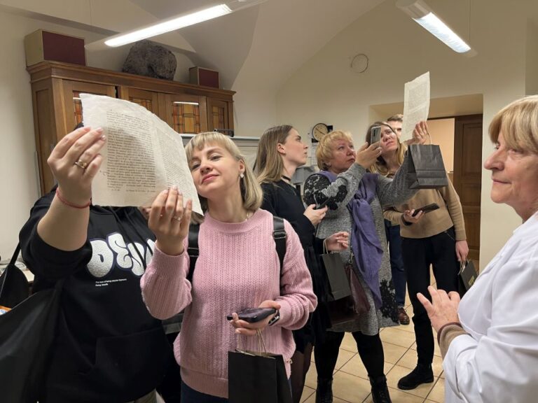 grupė žmonių apžiūrinėja rankose laikomus dokumentus ir fotografuoja juos patalpos fone