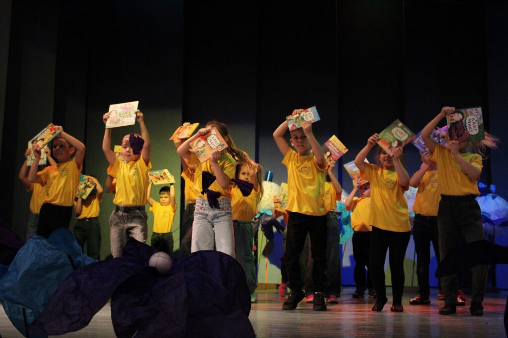 Nuotraukoje matoma grupė vaikų, kurie atlieka pasirodymą scenoje. Visi vaikai dėvi ryškiai geltonus marškinėlius ir laiko knygas virš galvų. Kai kurie vaikai stovi priešais kitus, o scena apšviesta spalvingais šviesų efektais. Fone matyti mėlyni ir purpūriniai audiniai, kurie papildo scenos estetiką.