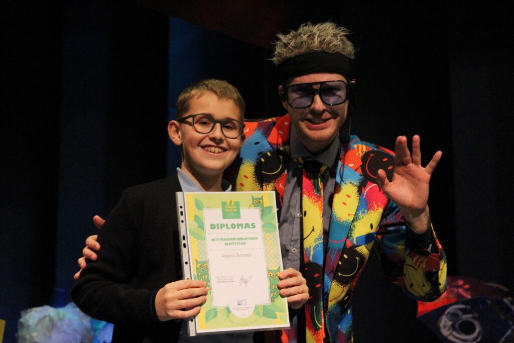 Nuotraukoje matyti berniukas ir žmogus su spalvotu kostiumu, stovintys scenoje. Berniukas šypsosi ir laiko diplomą, kuriame užrašyta „Diplomas". Jis dėvi akinius ir tamsų megztinį. Šalia jo stovi žmogus su ryškiai spalvotu kostiumu, dideliais akiniais ir mikrofonu, šypsodamasis ir keliantis ranką. Fone matyti dalis scenos dekoracijų.