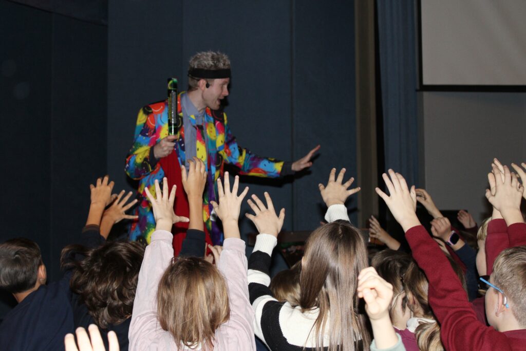 Nuotraukoje matyti spalvotai apsirengęs žmogus, stovintis scenoje ir rodančius ranką į priekį, tarsi bendraudamas su auditorija. Aplink jį yra vaikų rankos, ištiestos į viršų, tarsi jie laukia dėmesio ar nori užduoti klausimą. Spalvingas žmogaus kostiumas išsiskiria, o fone matosi tamsūs scenos kampai.