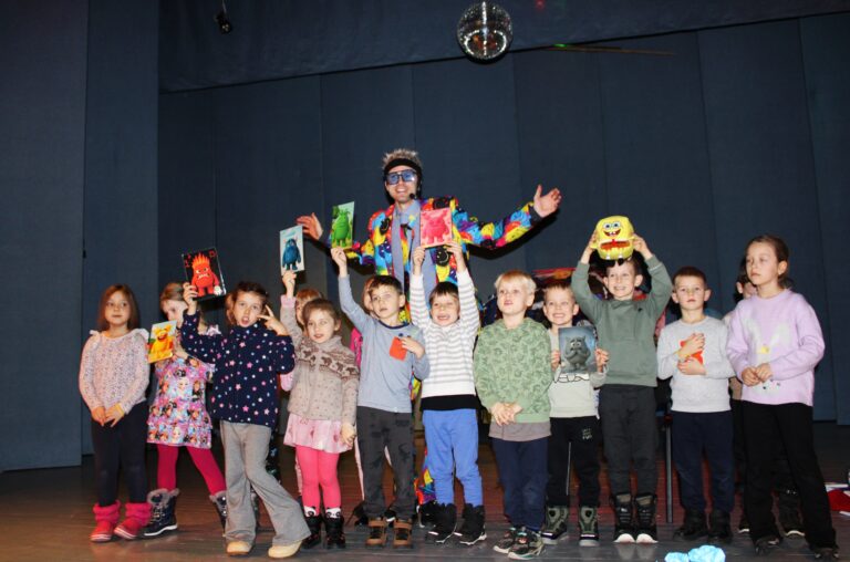 Scenoje stovi grupė vaikų kartu su spalvingai apsirengusiu artistas, vilkinčiu ryškų, margą kostiumą ir nešiojančiu didelius akinius. Vaikai šypsosi ir laiko pakėlę įvairiaspalvius personažų paveikslėlius bei žaislą. Fone – tamsiai mėlynos sienos ir pakabintas diskotekos kamuolys.