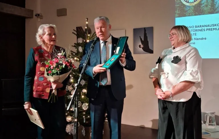 Nuotraukoje matomas apdovanojimo įteikimo momentas. Viduryje stovintis vyras laiko atidarytą apdovanojimo dėžutę, o šalia jo stovi dvi moterys. Pirmoji moteris laiko puokštę su gėlėmis ir žiūri į apdovanojimą, o antroji moteris stovi šalia su balta palaidine su juodais dekoro elementais. Fone matomas kalėdų eglutė su puošmenomis ir dekoracijomis.