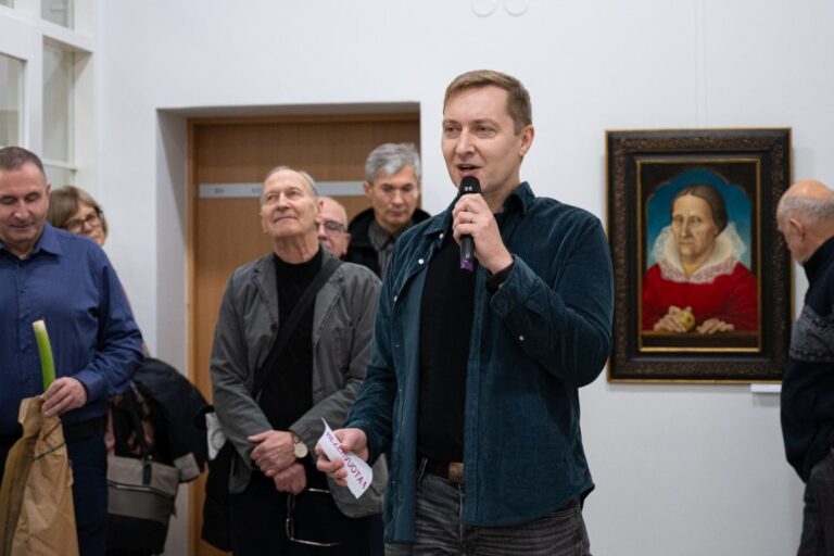 Vyras su mikrofonu stovi besiklausančių renginio dalyvių ir parodos eksponatų fone