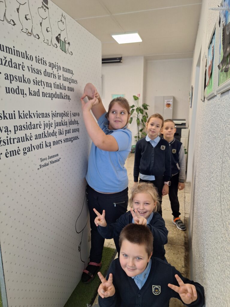 Nuotraukoje matyti grupė vaikų, kurie dalyvauja interaktyvioje veikloje su Tove Jansson knygos „Troliai Mumiai“ citata ir iliustracija. Vienas iš vaikų laikosi plakato, kuriame yra ištrauka iš knygos. Kiti vaikai stovintys eilėje priešais, šypsosi ir rodo „taikos“ ženklą. Plakate matosi troliai ir citata, skirta vaikams. Fone yra šviesus ir tvarkingas kambarys su kabinamais meno kūriniais ant sienos ir žaluma. Visi vaikai dėvi mokyklinius drabužius – mėlynus marškinėlius ir tamsias kelnes.