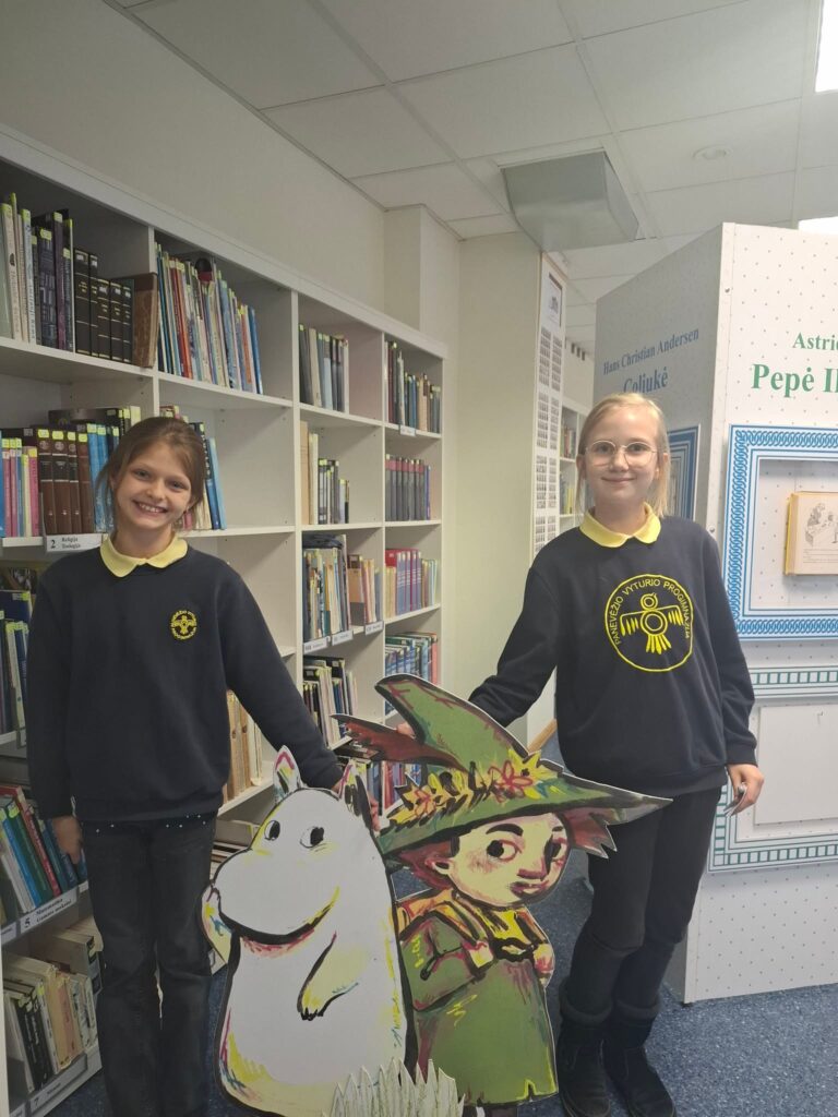 Nuotraukoje matomos dvi mergaitės, stovinčios prie spalvingo Muminų personažų stendo. Jos laikosi prie storo kartono figūrų – viena rodo į Muminą, o kita į Puką. Fone matoma biblioteka, kurioje už lentynų yra įvairių knygų. Mergaitės dėvi tamsiai mėlynus džemperius su geltonu Vyturio progimnazijos logotipu ir šviesiai geltonus marškinius po džemperiais.