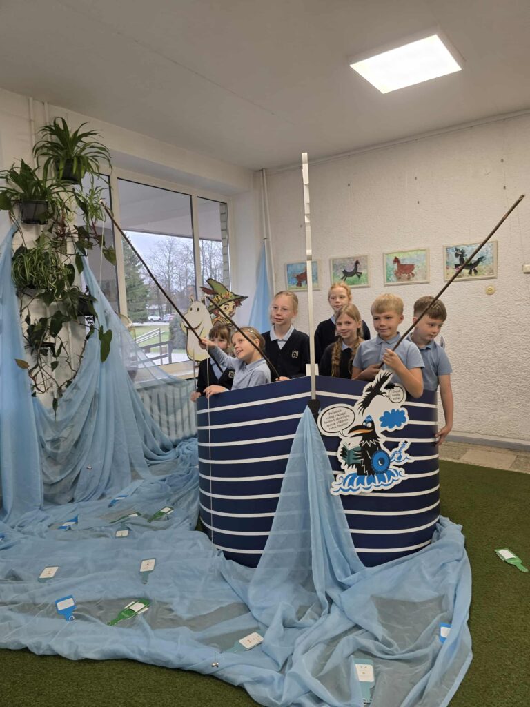 Nuotraukoje matyti grupė vaikų, kurie stovi ant improvizuoto laivo, kurio šonai pagaminti iš stilingų mėlynų ir baltų juostų. Vaikai žaidžia edukacinę veiklą, laikydami žvejybinio tipo meškeres ir interaktyviai bendraudami su aplinka. Laivas ir aplinka sukurtos iš mėlynos medžiagos, kuri simbolizuoja vandenį, o ant laivo matyti įvairūs personažai ir komiksų elementai. Fone matosi langas su žaliuojančia aplinka ir ant sienos kabančiais meno kūriniais, vaizduojančiais šunis.