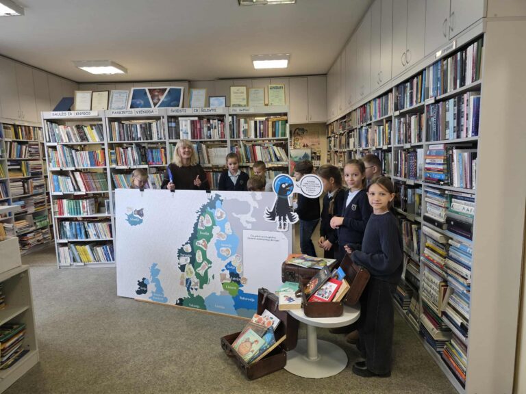 Nuotraukoje matome biblioteką, kurioje vaikai ir suaugusioji moteris dalyvauja edukacinėje veikloje. Moteris stovi šalia didelio žemėlapio, vaizduojančio Skandinavijos šalis ir kitas Europos šalis su įvairiais piešiniais. Vaikai stovi šalia jos, žiūri į žemėlapį ir kartu su ja dalyvauja veikloje. Žemėlapis atrodo kaip interaktyvus, jame matomi įvairūs gyvūnų atvaizdai, tikriausiai susiję su šalimi. Taip pat matyti stalas su atidarytomis lagaminų dėžėmis, kuriuose yra įvairių knygų. Bibliotekos aplinka yra šviesi ir pilna knygų, kurios yra tvarkingai sudėtos ant lentynų.